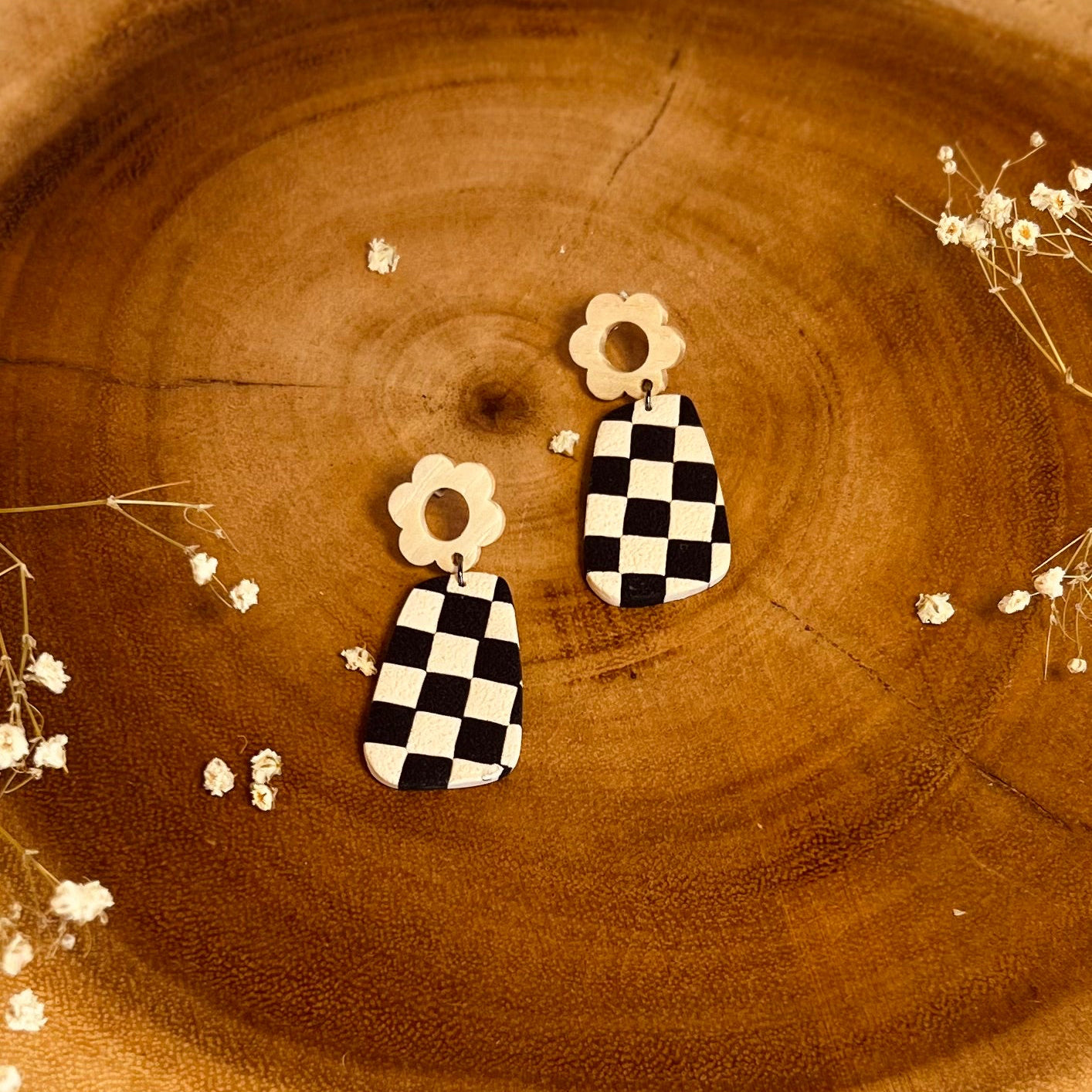 A close-up image of a pair of black and white checkered earrings placed on a wooden surface with natural wood flower accents.