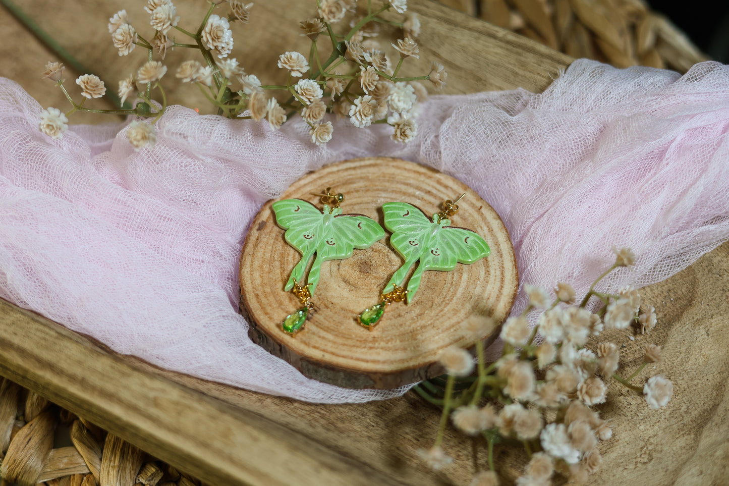 Wooden Coaster with shimmery green luna moth earrings with gold hardware on a floral background with tulle