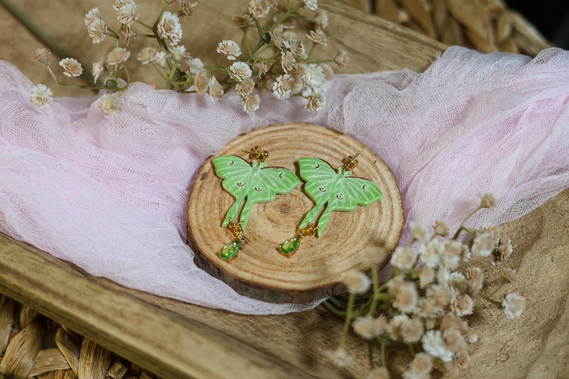 Wooden Coaster with shimmery green luna moth earrings with gold hardware on a floral background with tulle