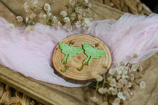 Wooden Coaster with shimmery green luna moth earrings with gold hardware on a floral background with tulle