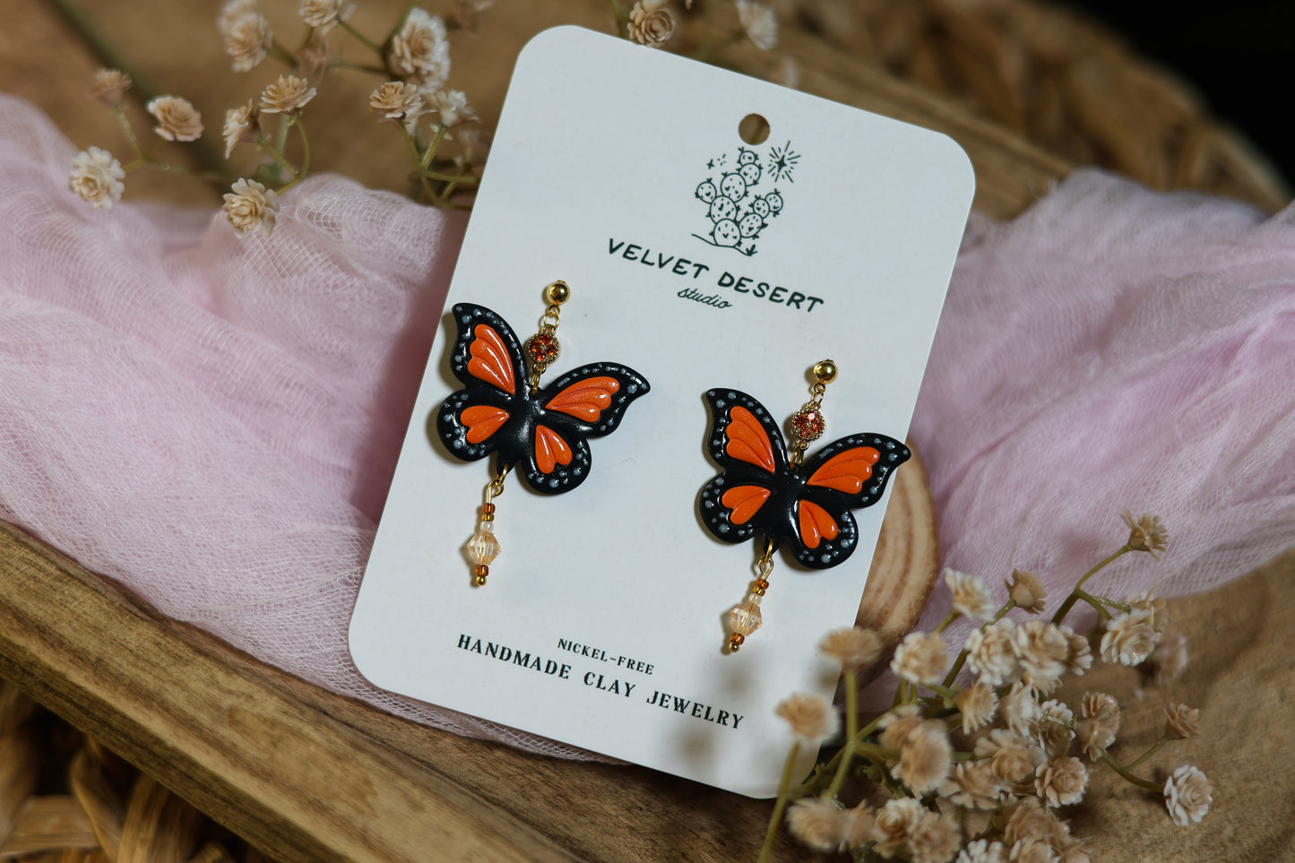 Butterfly-shaped earrings on a 'Velvet Desert' branded card with flowers in the background