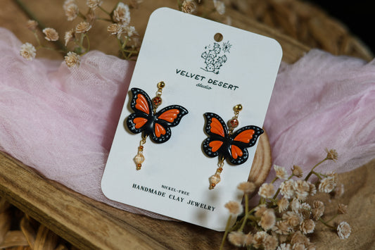 Butterfly-shaped earrings on a 'Velvet Desert' branded card with flowers in the background