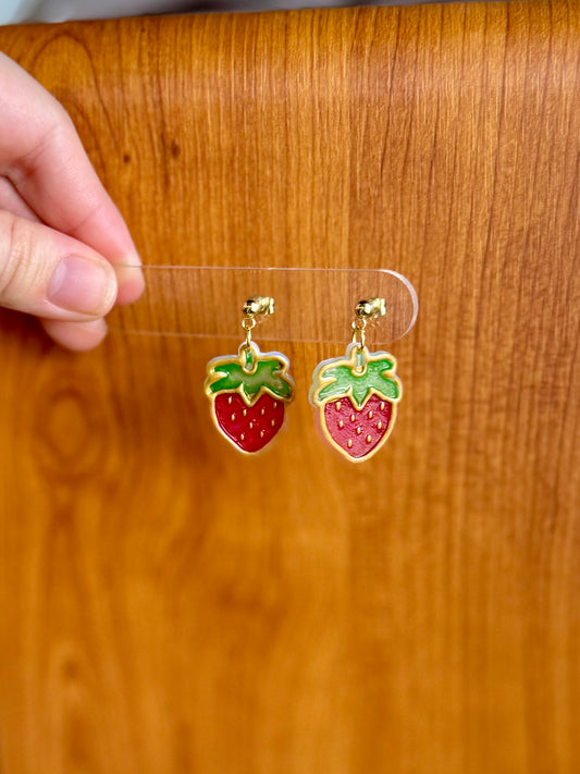 Strawberry-shaped earrings on a clear card held by a hand against a wooden background